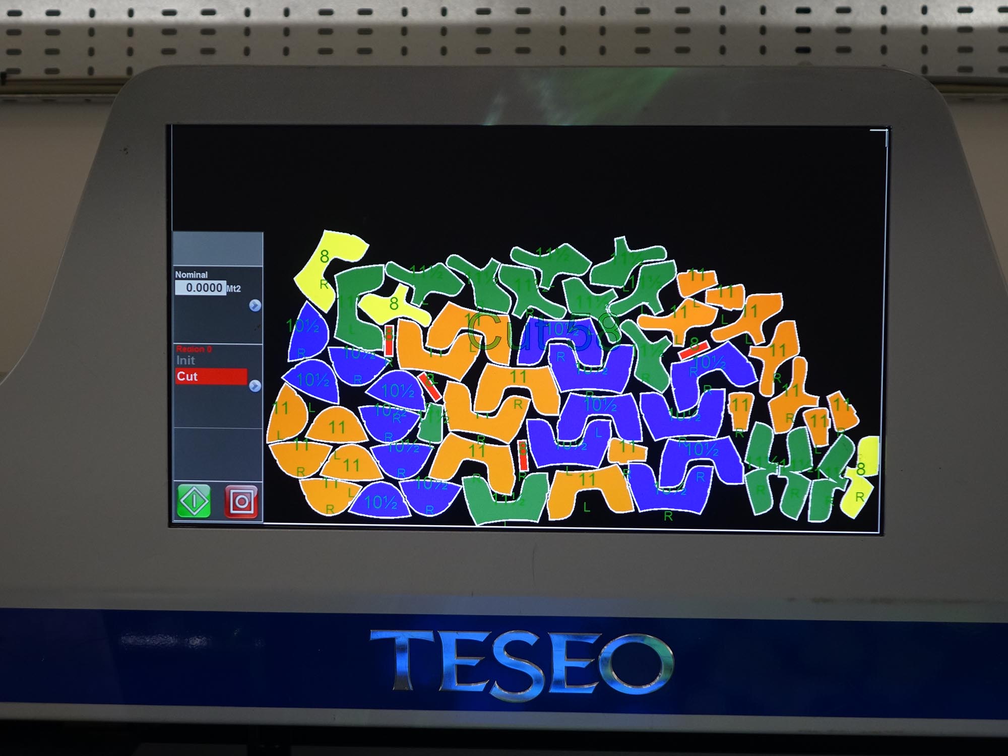 A digital screen displays a layout of multicoloured, numbered shapes for material cutting, arranged closely together on a black background. The interface shows controls and data on the left side. The machine is labelled TESEO.