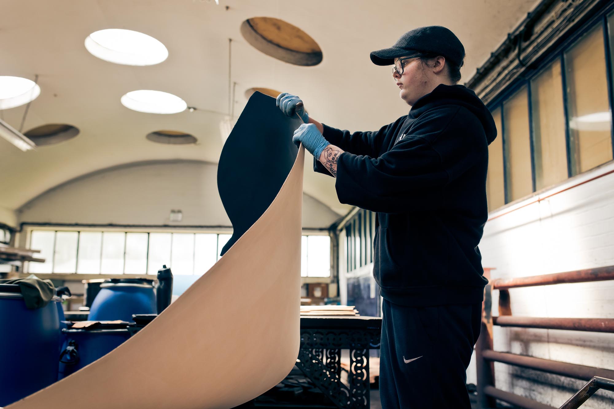 A person wearing glasses, a cap, and gloves inspects or lifts a large sheet of material in a workshop with circular skylights and barrels in the background.