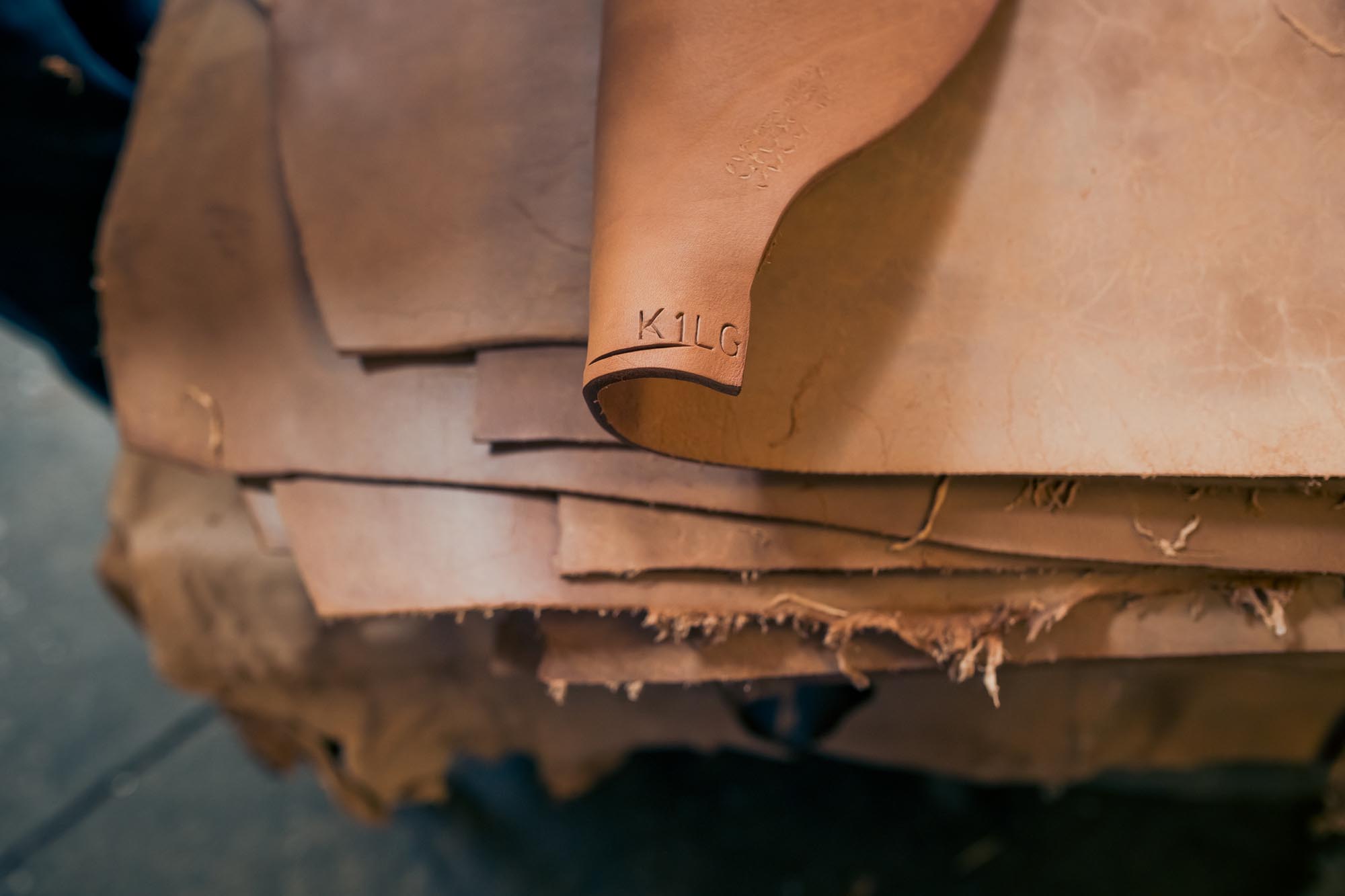 A stack of tanned leather sheets with one sheet curled at the corner, showing the embossed text KILG. The edges of the leather are uneven and rough.