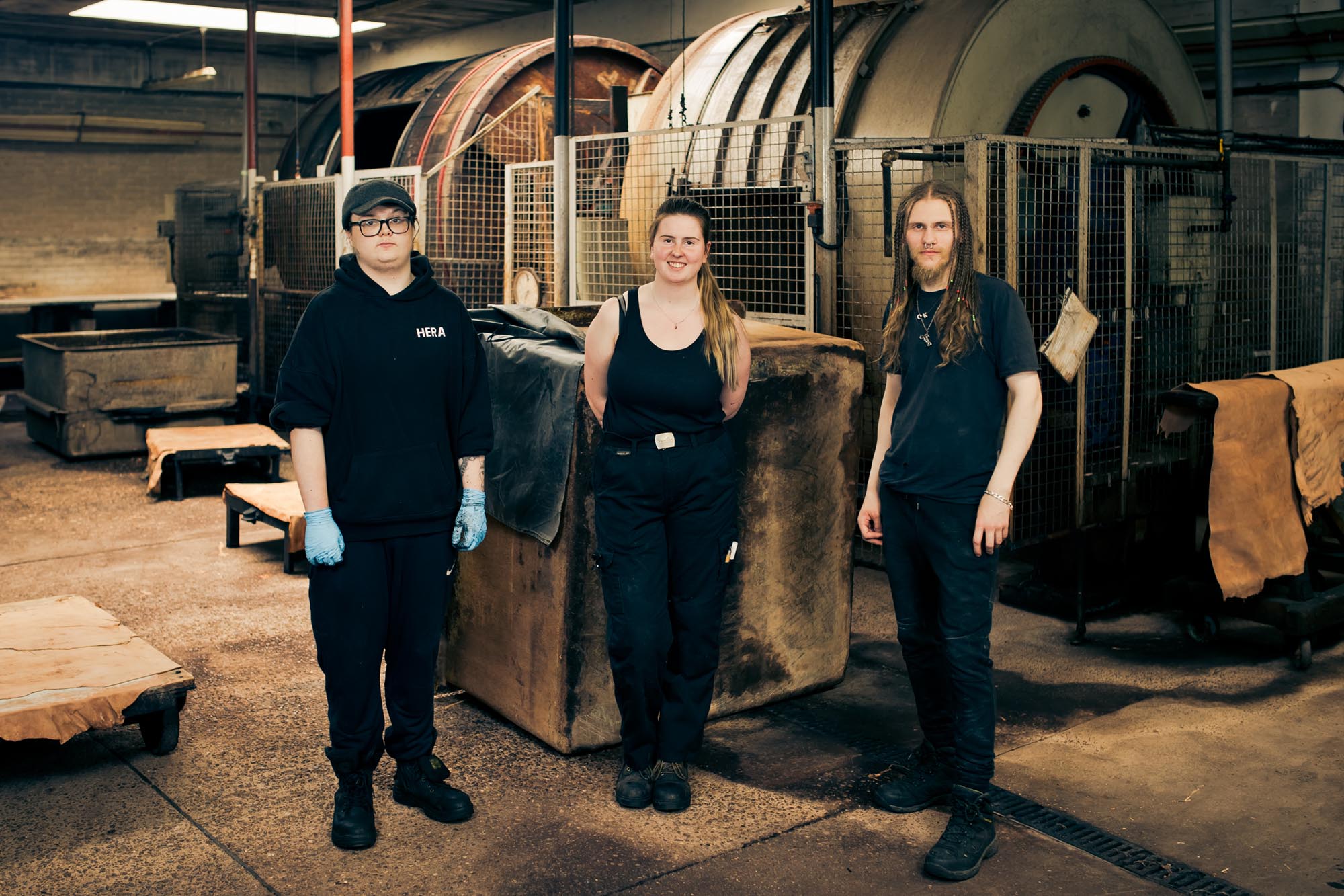 Three people stand in an industrial workshop with large machinery and equipment in the background. They wear dark clothing and work gloves, and some hides or leather pieces are visible around them.