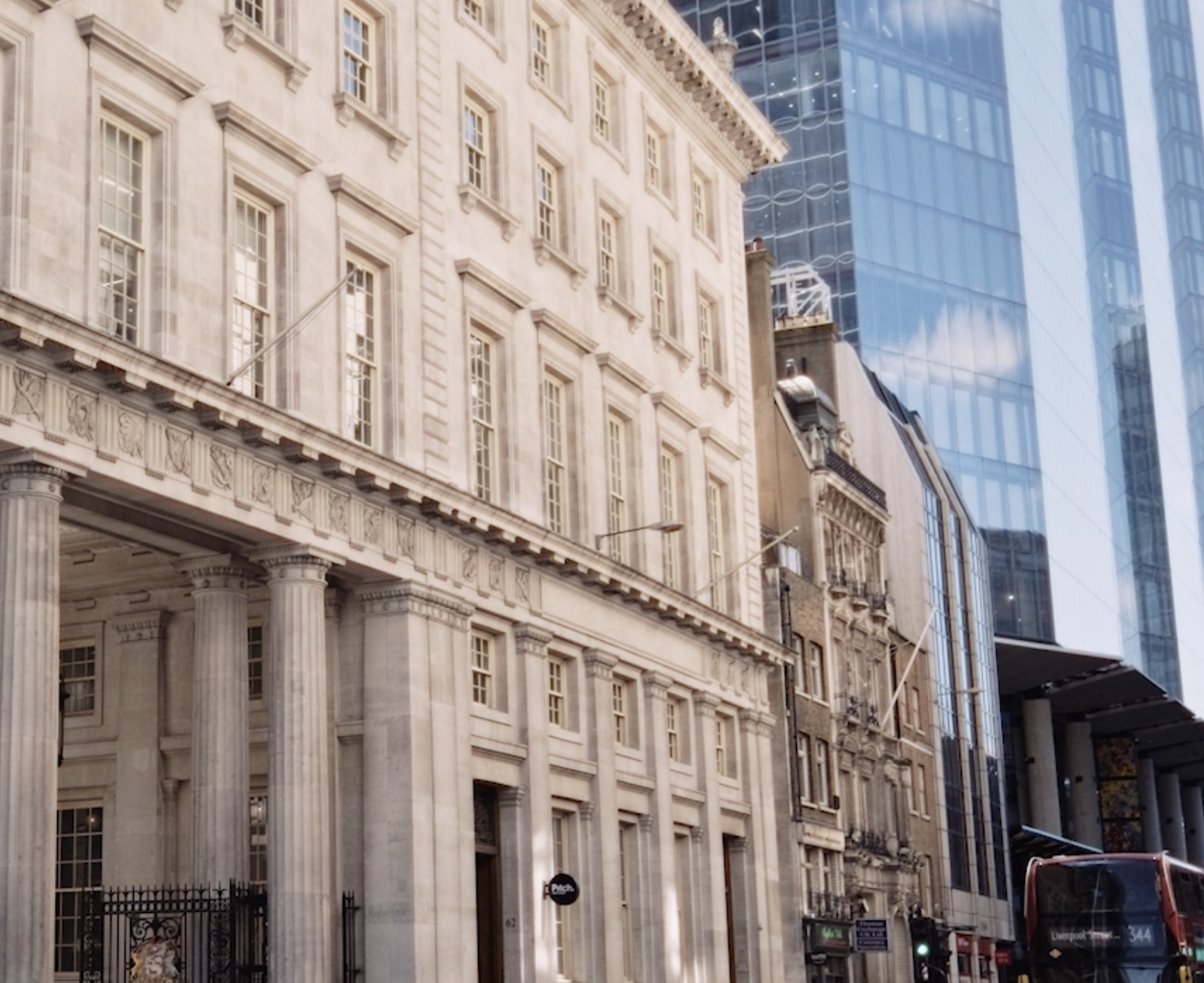 A neoclassical stone building with tall columns stands next to modern glass skyscrapers along a city street. A red double-decker bus is visible in the lower right corner.