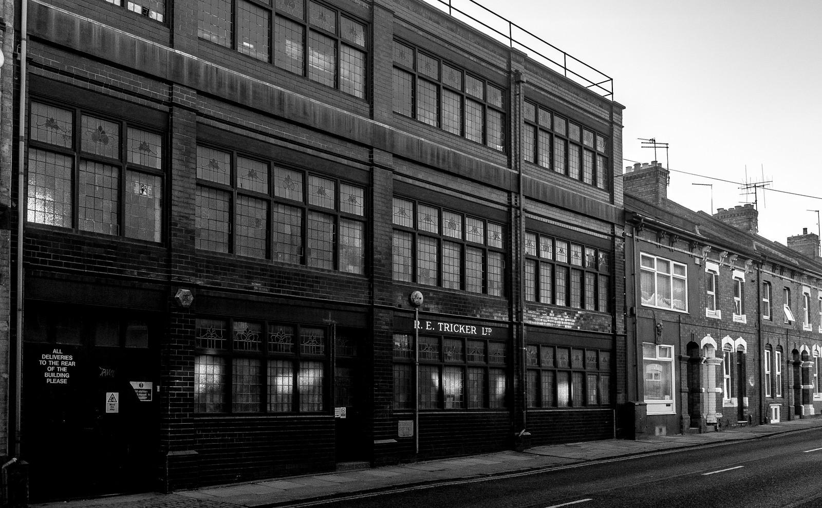 A black and white photo of an empty street with brick buildings. One building has large multi-pane windows and a sign that reads R. E. Tricker Ltd. The street appears quiet with no people or vehicles visible.