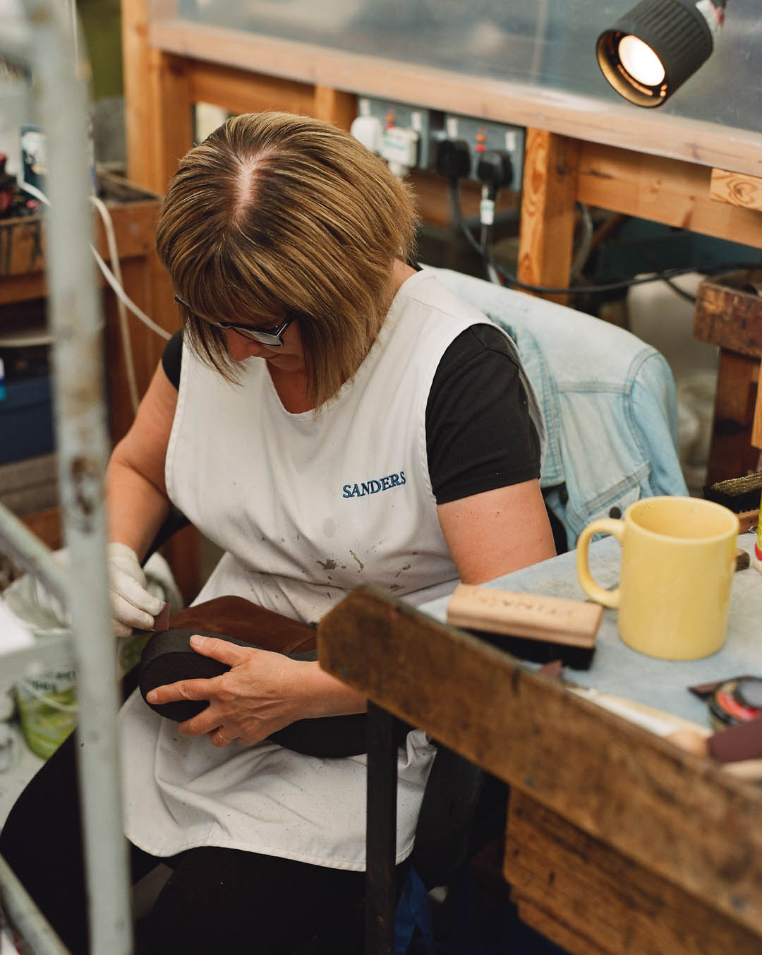 A woman in a white apron works on a shoe at a workbench next to a yellow mug and lamp.