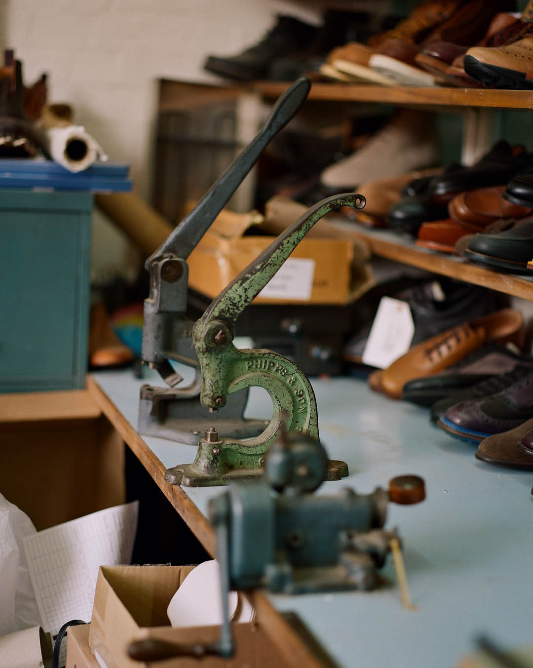 A green metal hand press sits on a workbench in a workshop, surrounded by shoe repair equipment and shelves filled with various pairs of shoes.