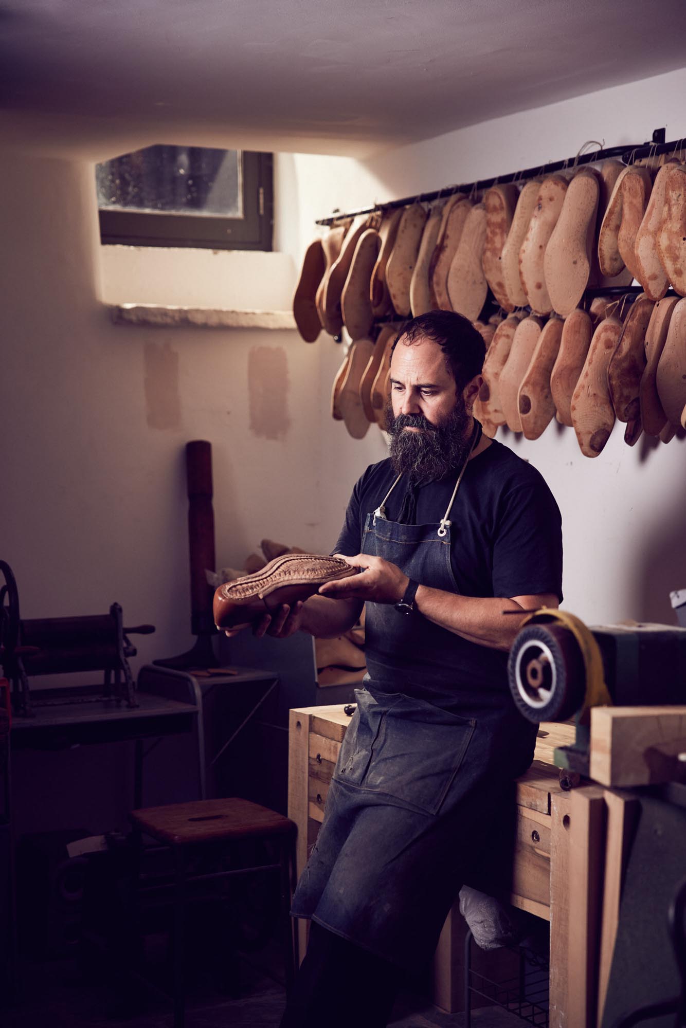 A bearded man wearing an apron sits on a workbench in a workshop, examining a hand-crafted leather shoe sole. Leather shoe soles hang on the wall behind him, and tools are visible around the workspace.