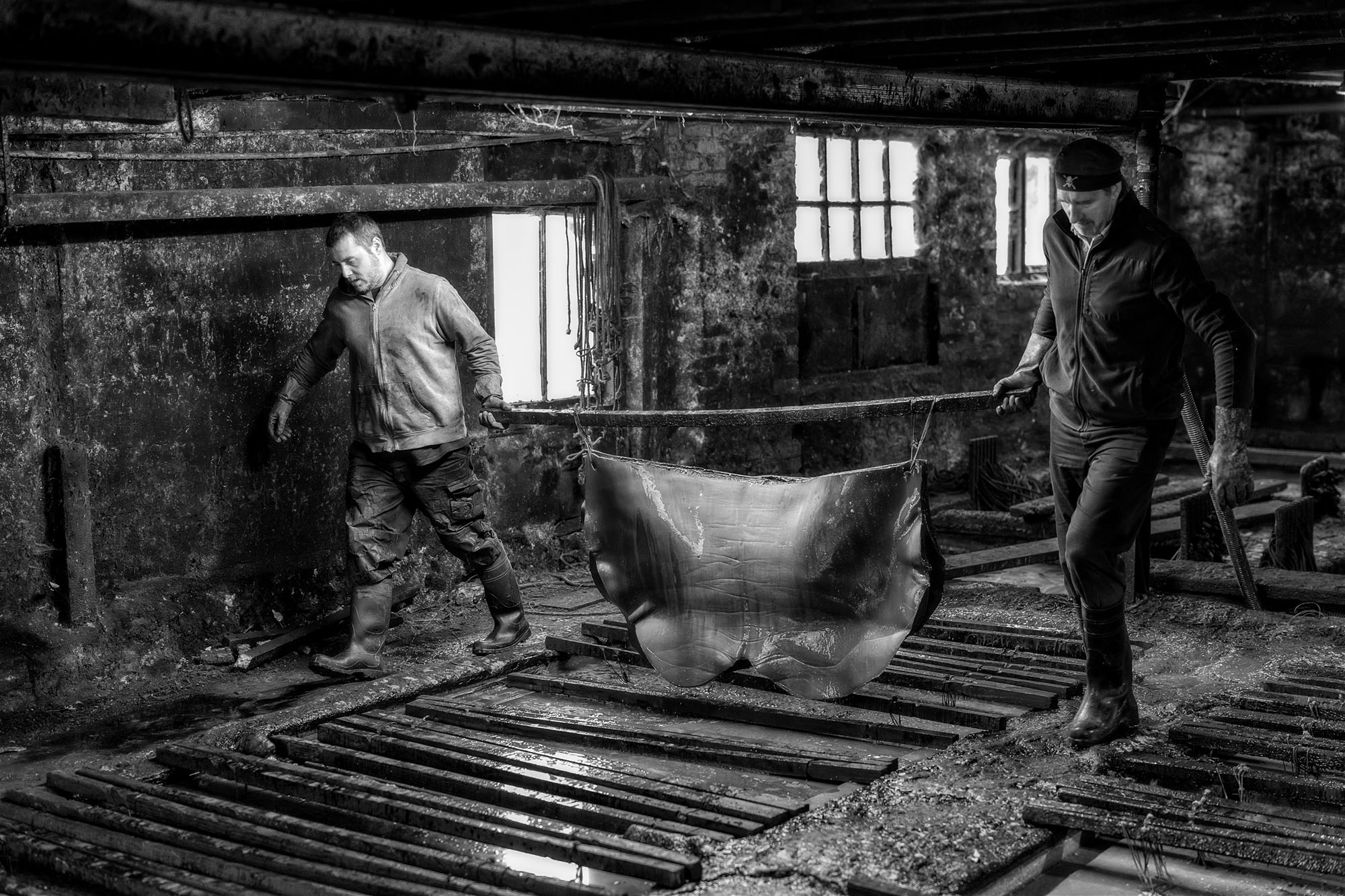Two men in work clothes carry a large piece of animal hide between wooden vats inside a dim, industrial workshop with rough walls and barred windows. The image is in black and white.