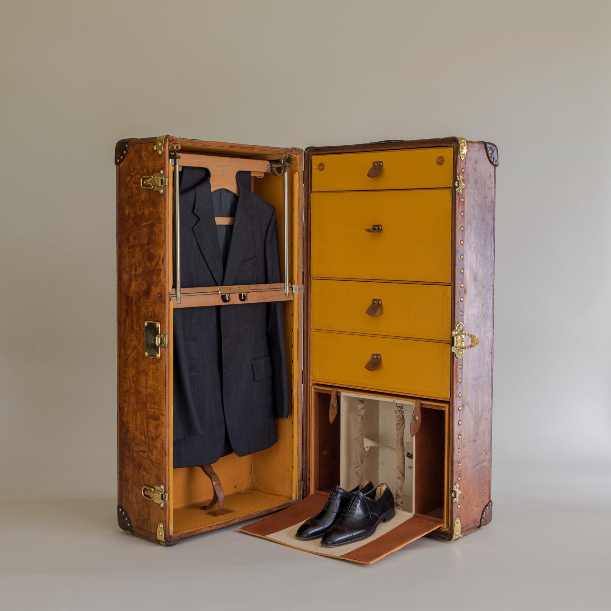A vintage brown leather trunk is open, revealing a hanging dark suit, four yellow drawers, two pairs of black shoes, and compartments holding two pairs of trousers. The trunk stands on a neutral background.