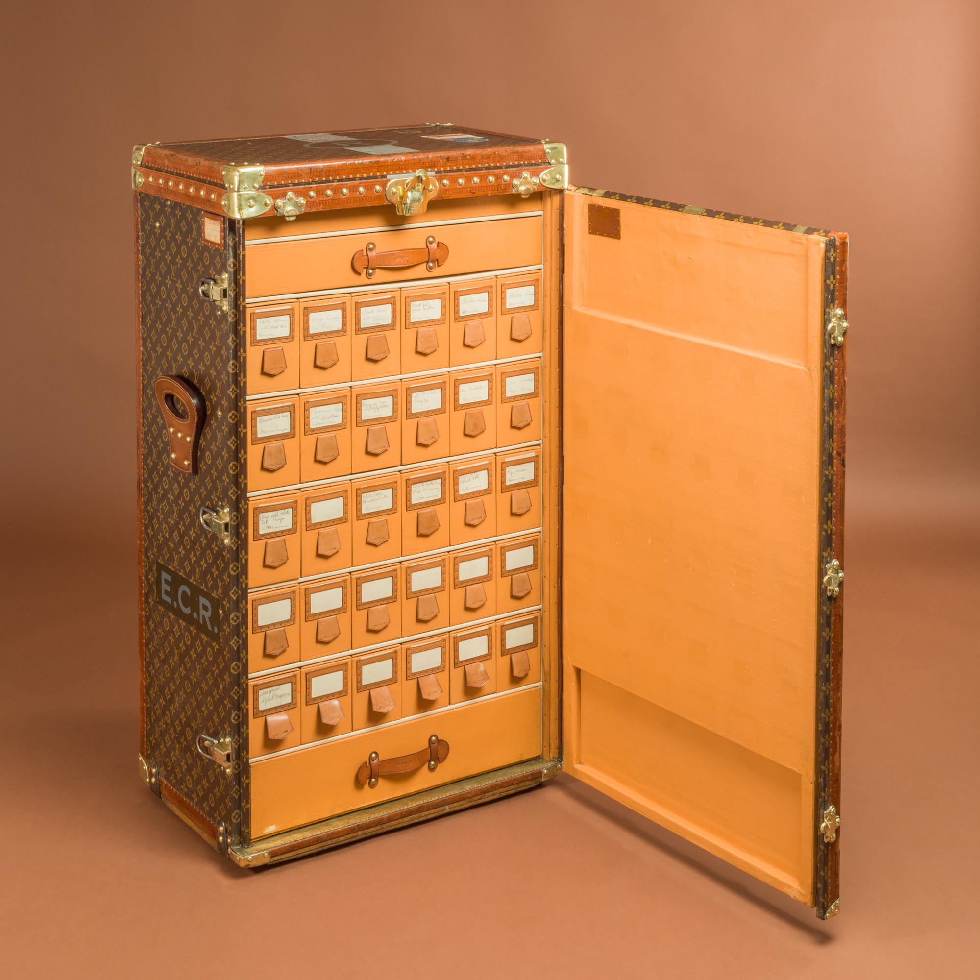 A vintage Louis Vuitton trunk is open to reveal a set of 20 labelled drawers arranged in four rows. The exterior is brown with a patterned design, whilst the interior features tan leather. The initials E.C.R. are on the side.