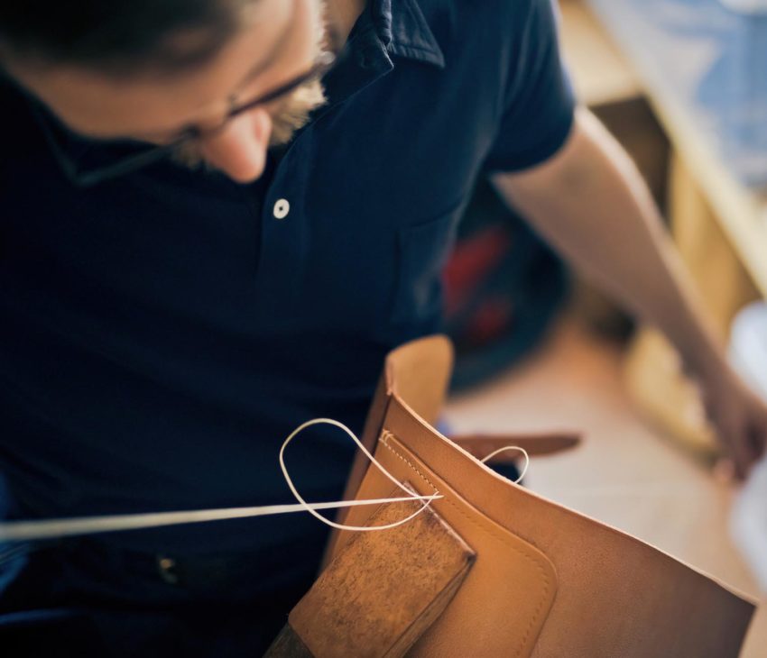 A person hand-stitching leather with white thread, focussing intently on their detailed craftsmanship.