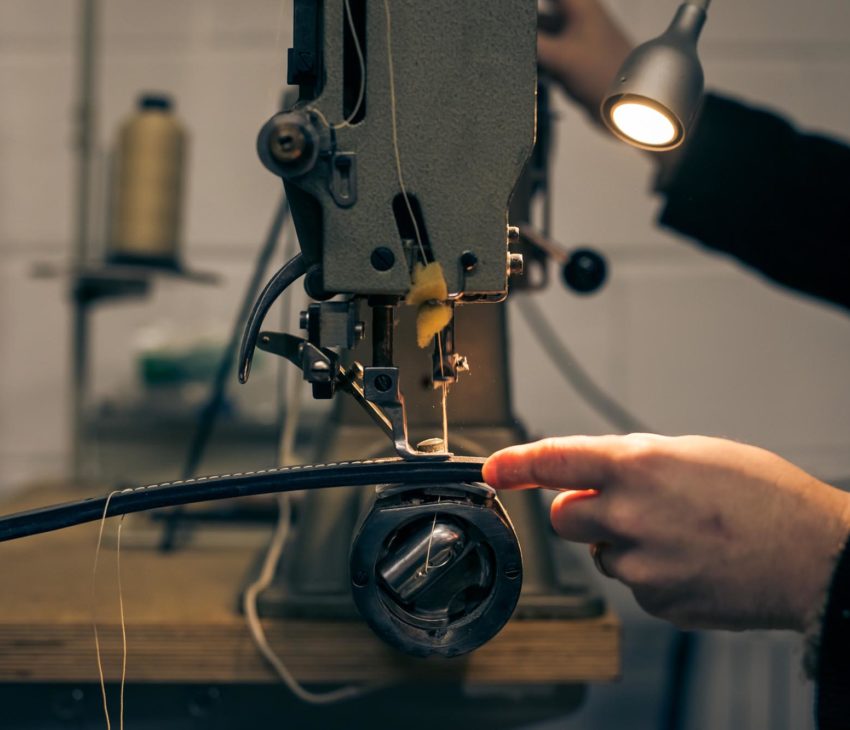 A close-up of a person’s hands using an industrial sewing machine to stitch a piece of black material, with a spool of thread visible in the background and a small lamp illuminating the work area.