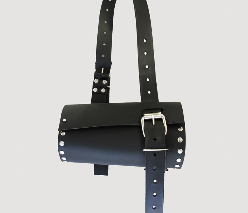 A black cylindrical leather bag with metal studs and a buckle, featuring an adjustable strap and an elongated fastening strap, displayed against a plain light grey background.