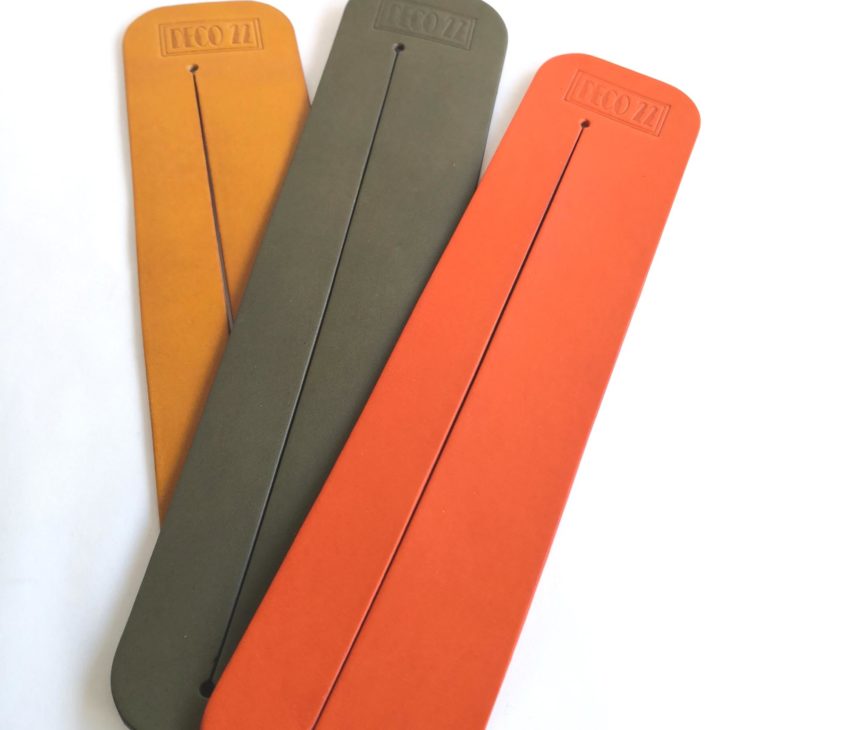 Three rectangular, flat leather bookmarks in orange, olive green, and mustard yellow colours, each with a single vertical slit down the centre and the word ECOZZI embossed at the top, displayed on a white background.