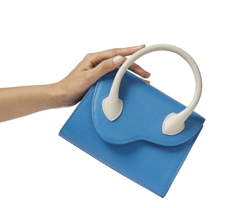 A hand holding a small blue handbag with a textured surface and two curved white handles against a white background.