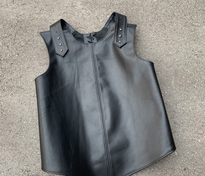 A sleeveless black leather waistcoat with a central seam, decorative stitching, and metal rivets on the shoulder straps, displayed on a rough grey concrete surface.