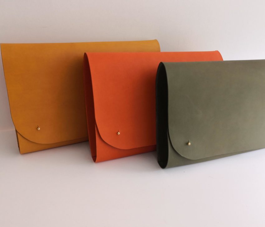 Three slim, rectangular envelope-style leather folders are displayed upright in a row. The folders are yellow, orange, and olive green, each with a single gold button fastening on the front. The background is plain white.