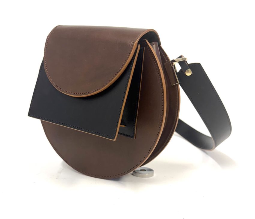 A round brown leather crossbody bag with a black front flap and black adjustable strap, placed upright on a white background.