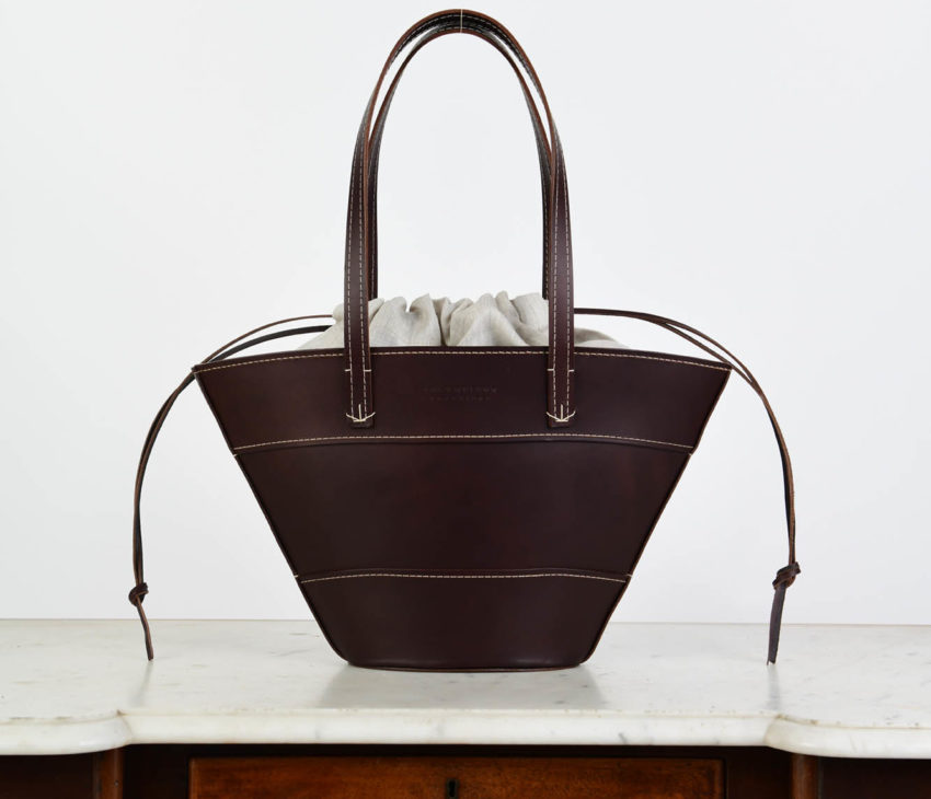 A dark brown leather handbag with two handles and a drawstring fabric insert is placed on a marble-topped wooden table with a drawer, against a plain white background.