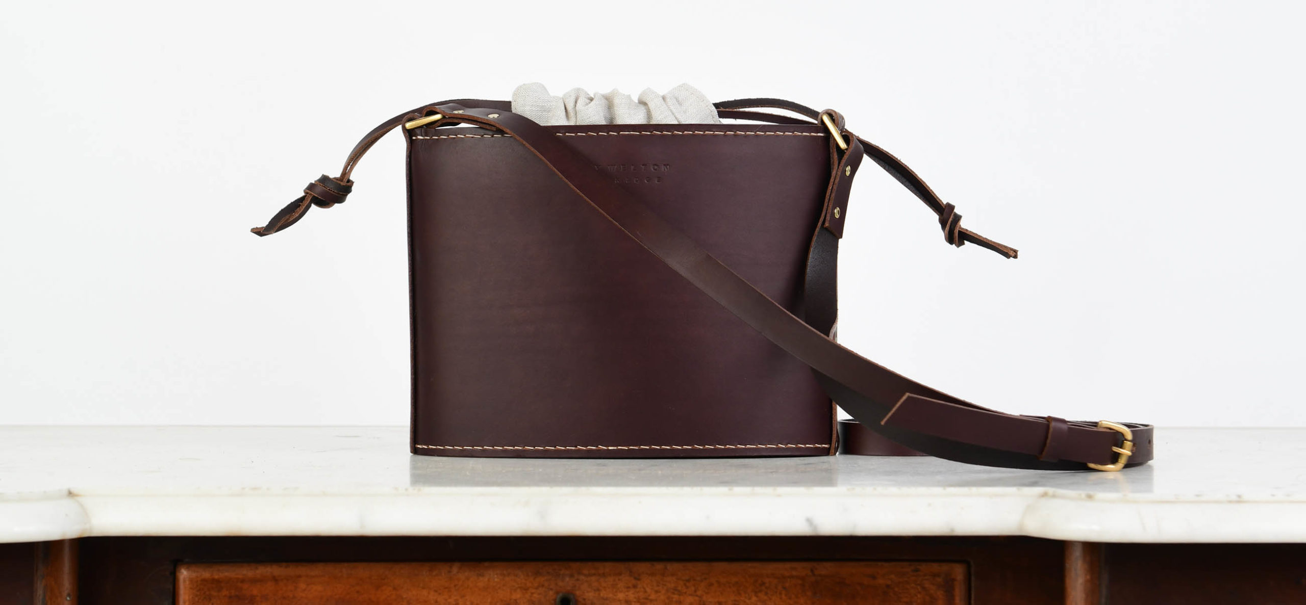 A dark brown leather crossbody bag with a wide adjustable strap, gold hardware, and a light fabric drawstring fastening sits on a white marble surface above a wooden drawer.