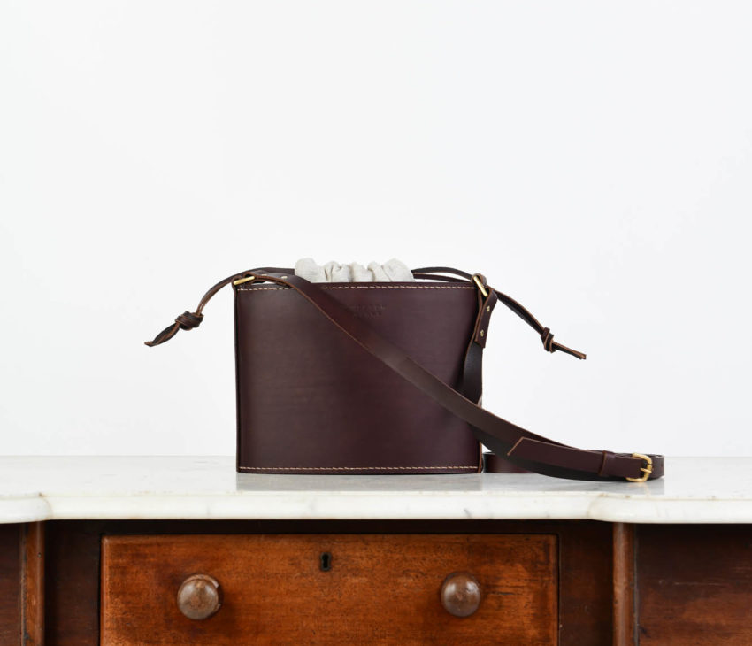 A dark brown leather crossbody bag with a long strap sits on a wooden desk with a marble top and two drawers against a plain white background.