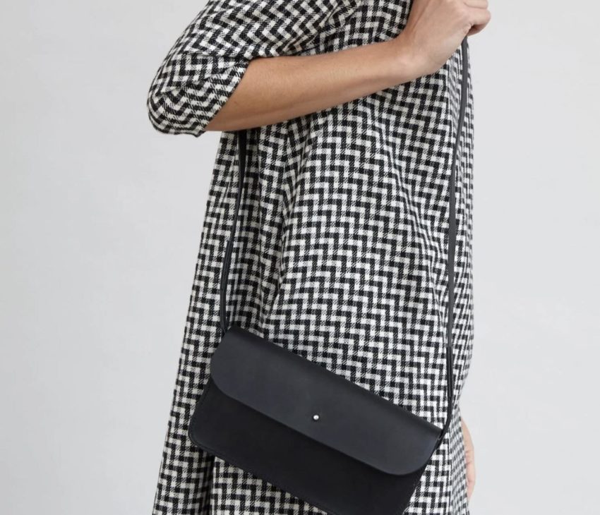 A person wearing a black-and-white houndstooth-patterned dress is shown from the neck down, carrying a black crossbody bag with a flap fastening and thin strap over their shoulder.