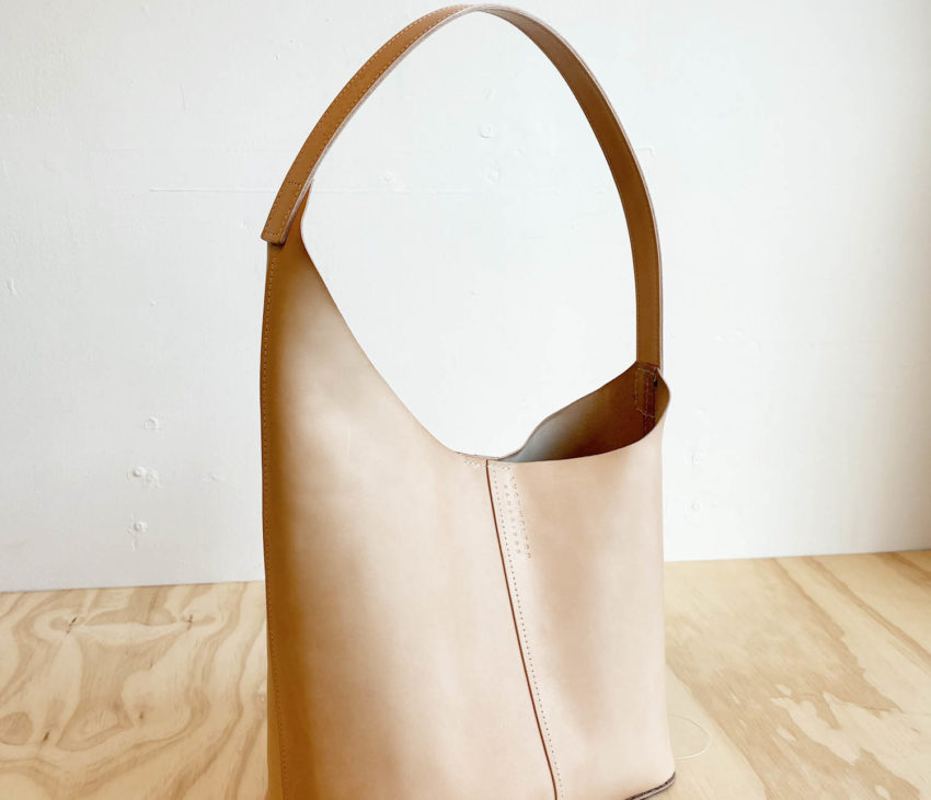 A light tan leather tote bag with a single wide strap, standing upright on a wooden surface against a plain, off-white background. The bag has clean lines and a minimalist design.